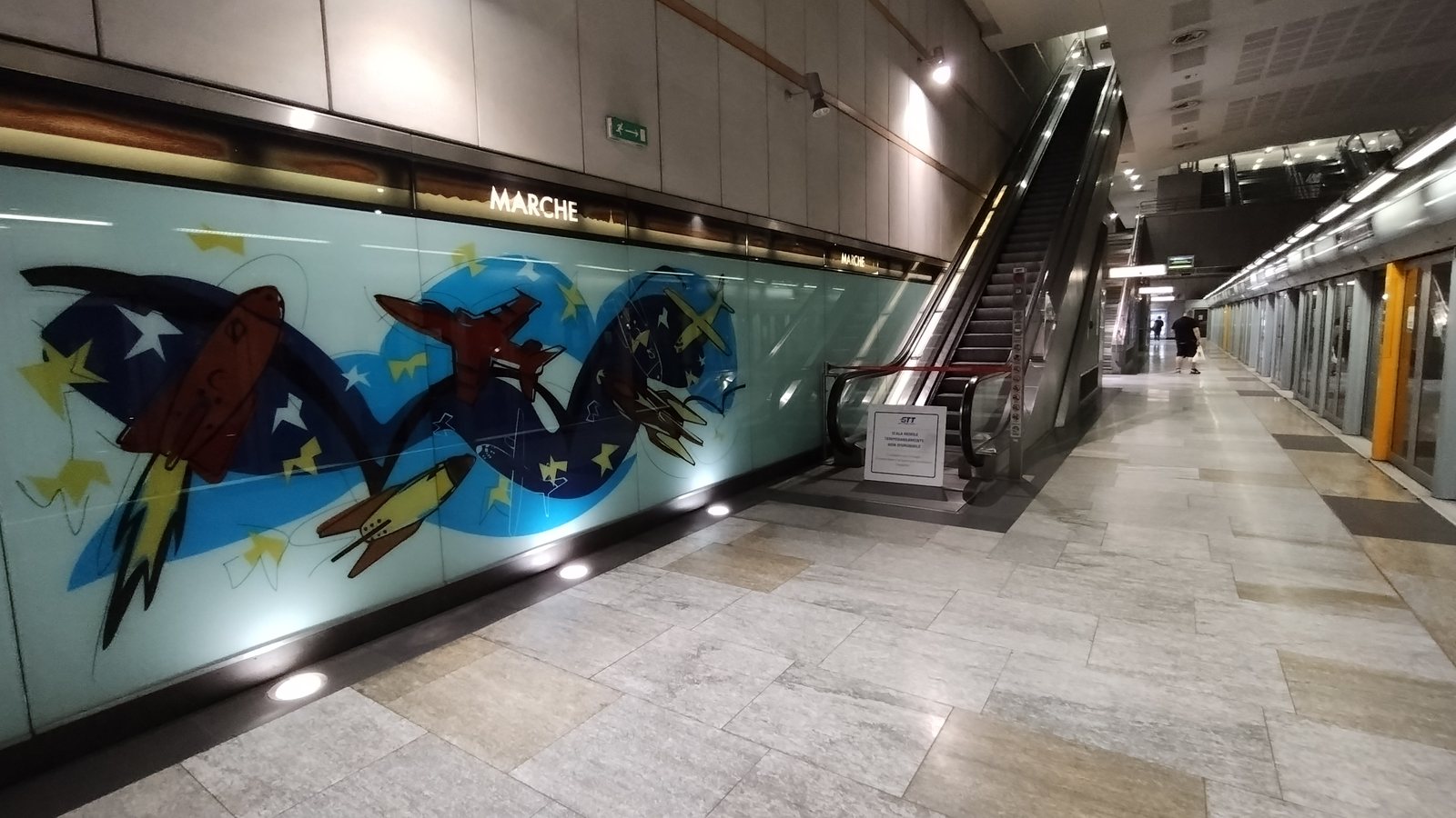 Banchina della stazione Marche: targa MARCHE, parete decorata e treno dietro le porte di banchina